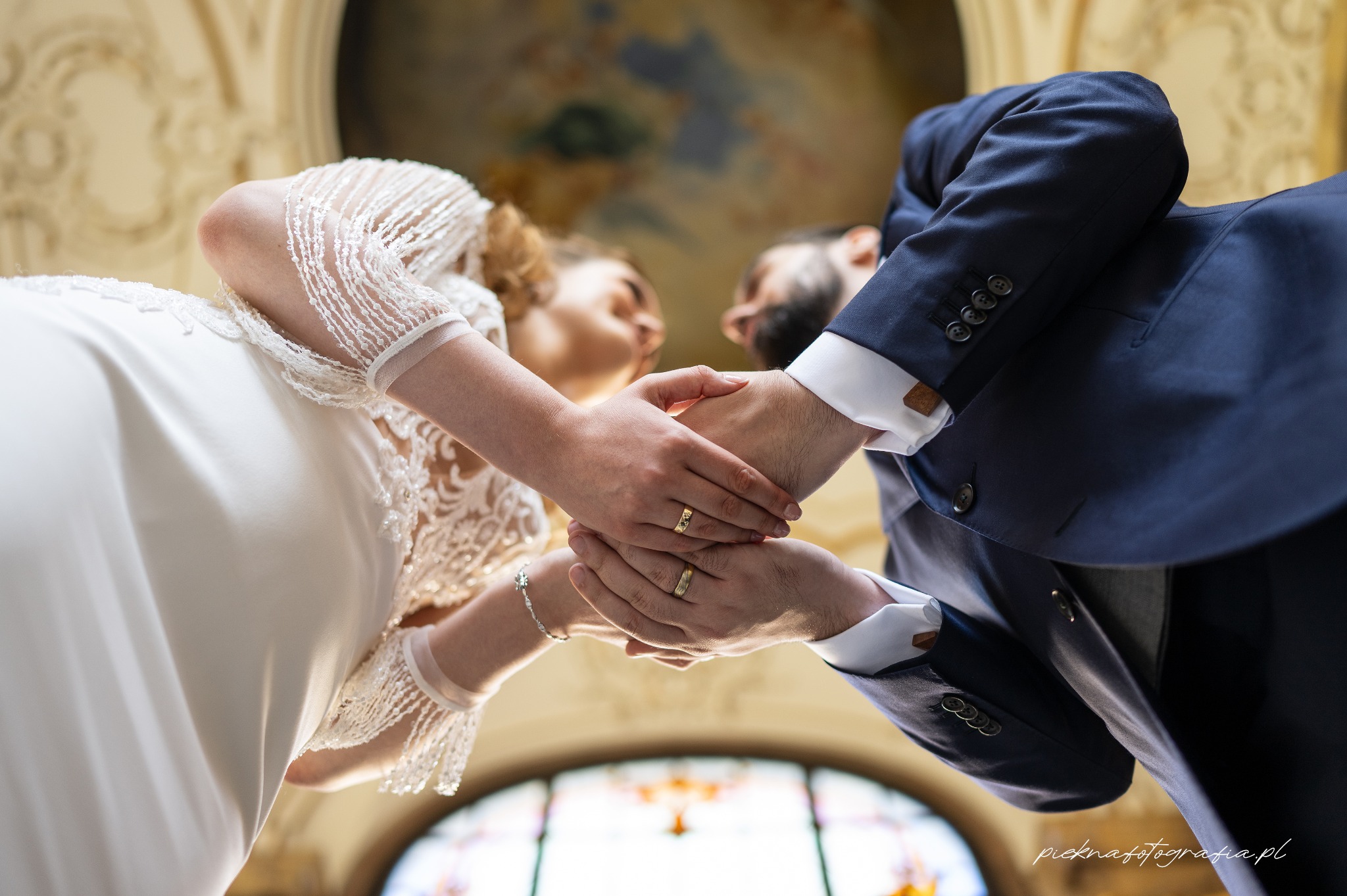 Nowożeńcy trzymają się za ręce, widoczne złote obrączki, suknia ślubna z koronkowymi rękawami, ciemny garnitur, widok z dołu na ozdobny sufit.