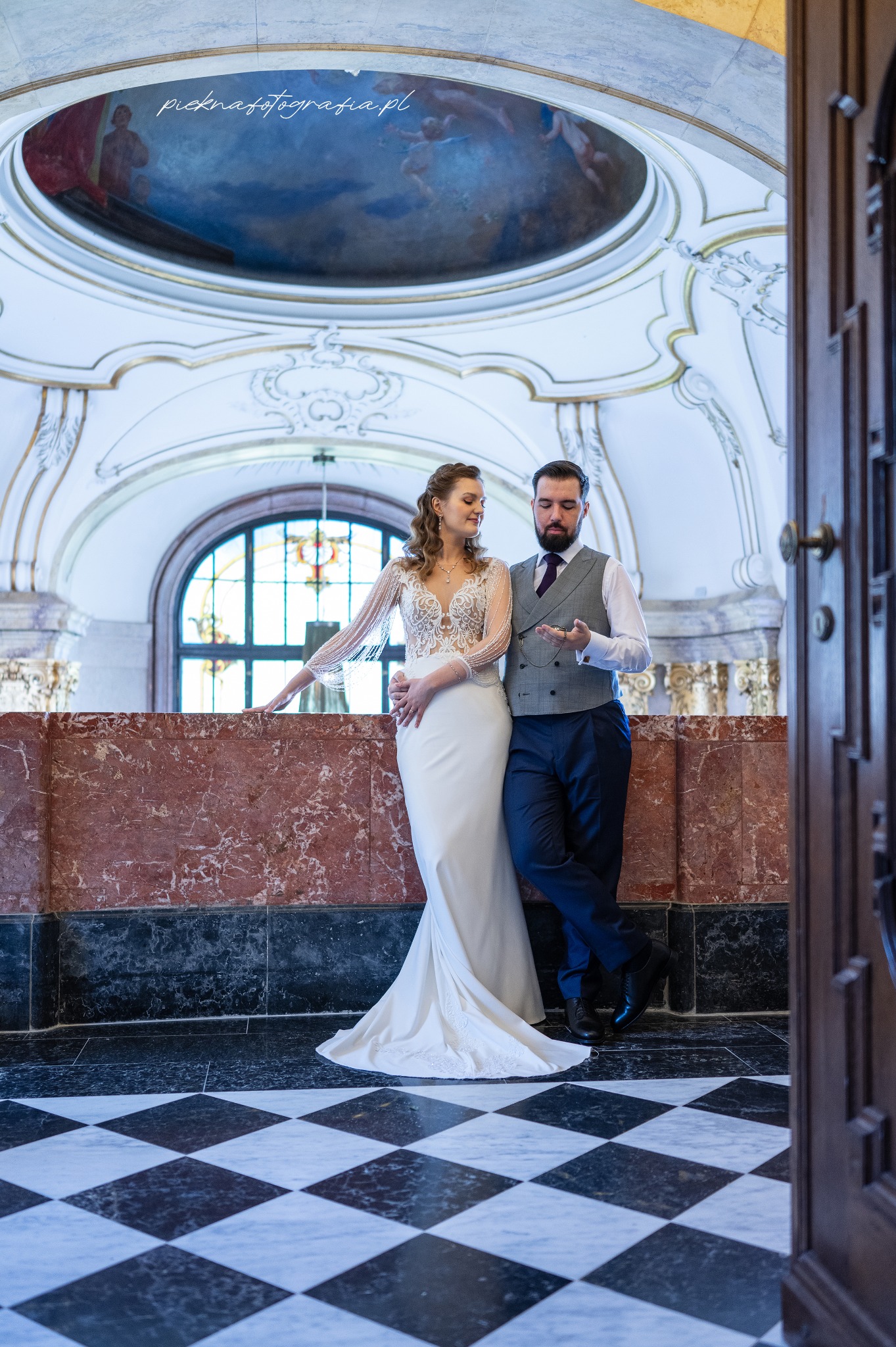 Elegancka para młoda w zabytkowym wnętrzu: panna młoda w koronkowej sukni z długimi rękawami, pan młody w kamizelce i z zegarkiem kieszonkowym, marmurowe ściany i posadzka w szachownicę.