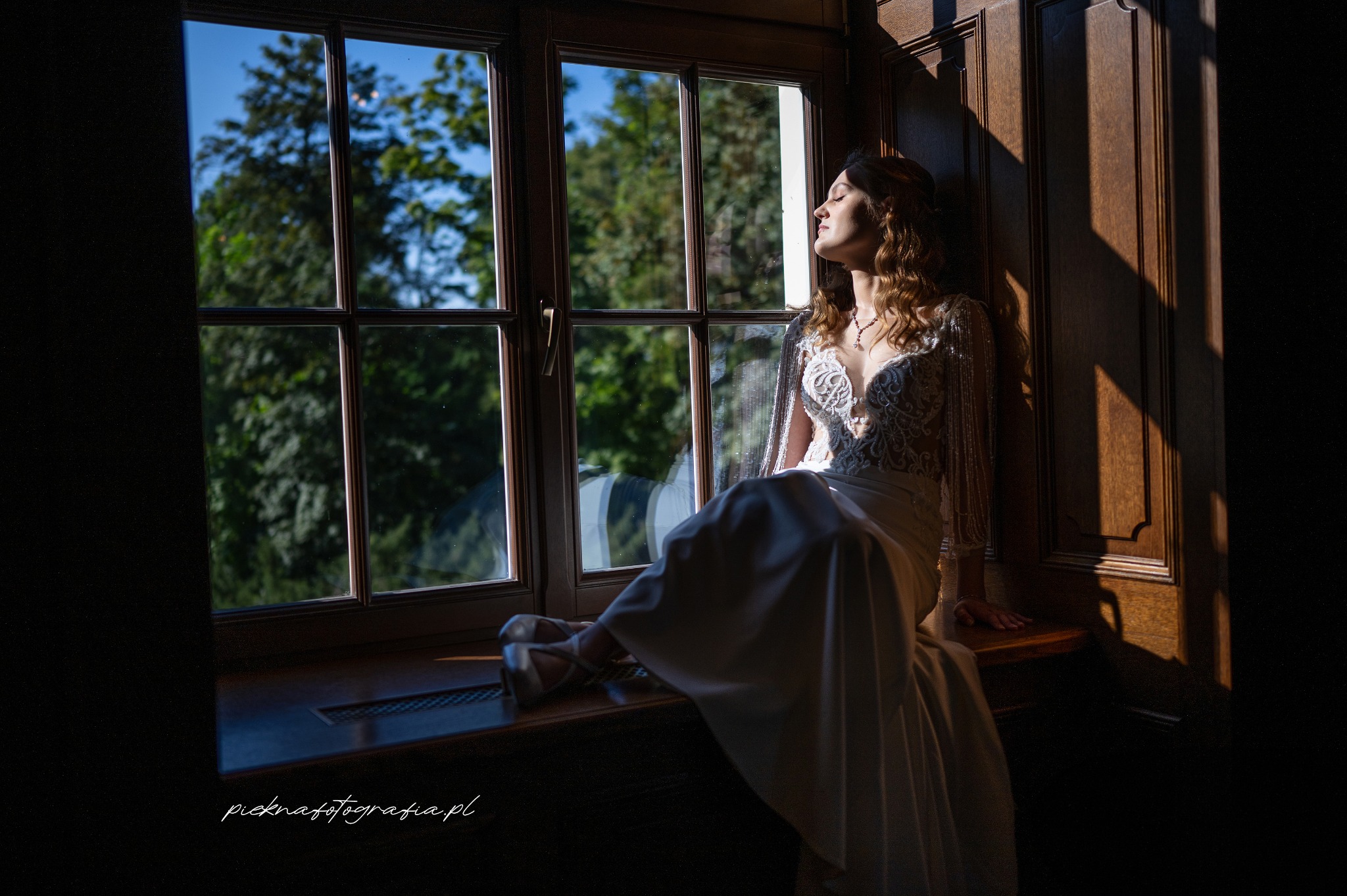 Panna młoda w koronkowej sukni siedzi na parapecie okna, twarz skierowana ku słońcu, w tle widok na zielone drzewa.