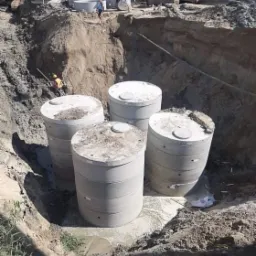 Cztery betonowe zbiorniki umieszczone w głębokim wykopie, widoczny pracownik schodzący po drabinie, w tle koparka i inne maszyny budowlane.