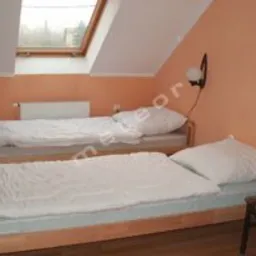 Pokój na poddaszu z dwoma pojedynczymi łóżkami z pościelą w odcieniach bieli, pomarańczowe ściany, okno dachowe i lampa ścienna. Widoczny fragment krzesła i podłogi z paneli.