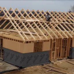 Konstrukcja drewniana domu w trakcie budowy z widoczną więźbą dachową i pracownikiem na dachu, zabezpieczona folią fundamentowa z nadrukiem, drabina oparta o ścianę.