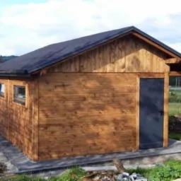 Mały, parterowy budynek z drewna, z czarnym dachem i drzwiami, na betonowej podstawie, z widocznym krajobrazem w tle.