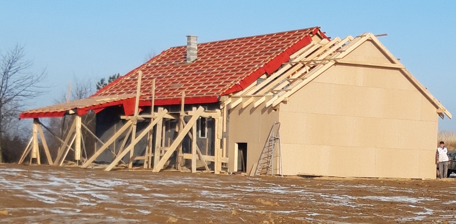 Budowa domu: widok na częściowo ukończony budynek z czerwoną dachówką, fragmentem więźby dachowej, zewnętrzną ścianą z płyt OSB oraz rusztowaniami z drewnianych bali. Na pierwszym planie błotniste...