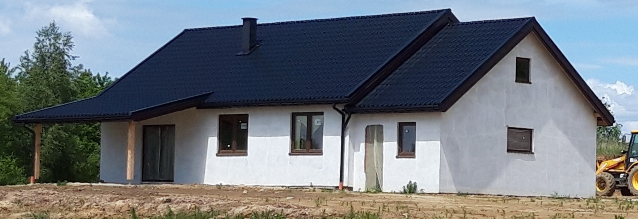Niedokończony dom z białą elewacją i ciemnym dachem, widoczne okna z brązowymi ramami, elementy konstrukcyjne ganku i traktor w tle.