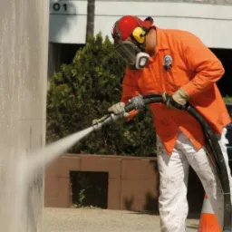 Pracownik w pomarańczowej koszuli i masce ochronnej czyści elewację budynku strumieniem wody pod ciśnieniem, widoczny efekt czyszczenia.