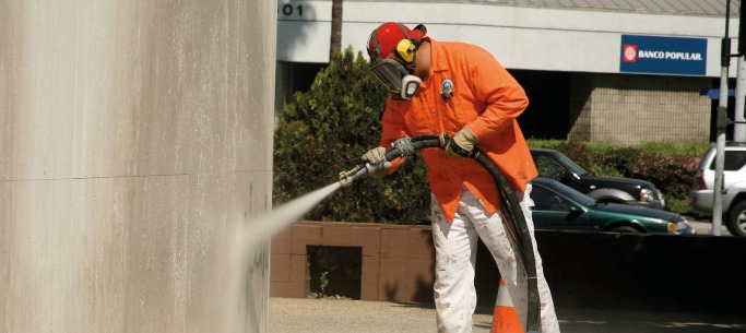 Pracownik w pomarańczowej koszuli i masce ochronnej czyści elewację budynku strumieniem wody pod ciśnieniem, widoczny efekt czyszczenia.