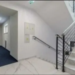 Wnętrze klatki schodowej w budynku biurowym z długim korytarzem w tle. Białe marmurowe schody i podłoga, ciemnoniebieska wykładzina na korytarzu, białe ściany z dwoma skrzynkami elektrycznymi.