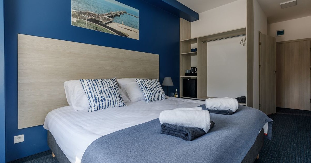 Pokój hotelowy z łóżkiem, szafą, minibarem i obrazem morza nad wezgłowiem, przygotowany dla gości z ręcznikami ułożonymi na łóżku.