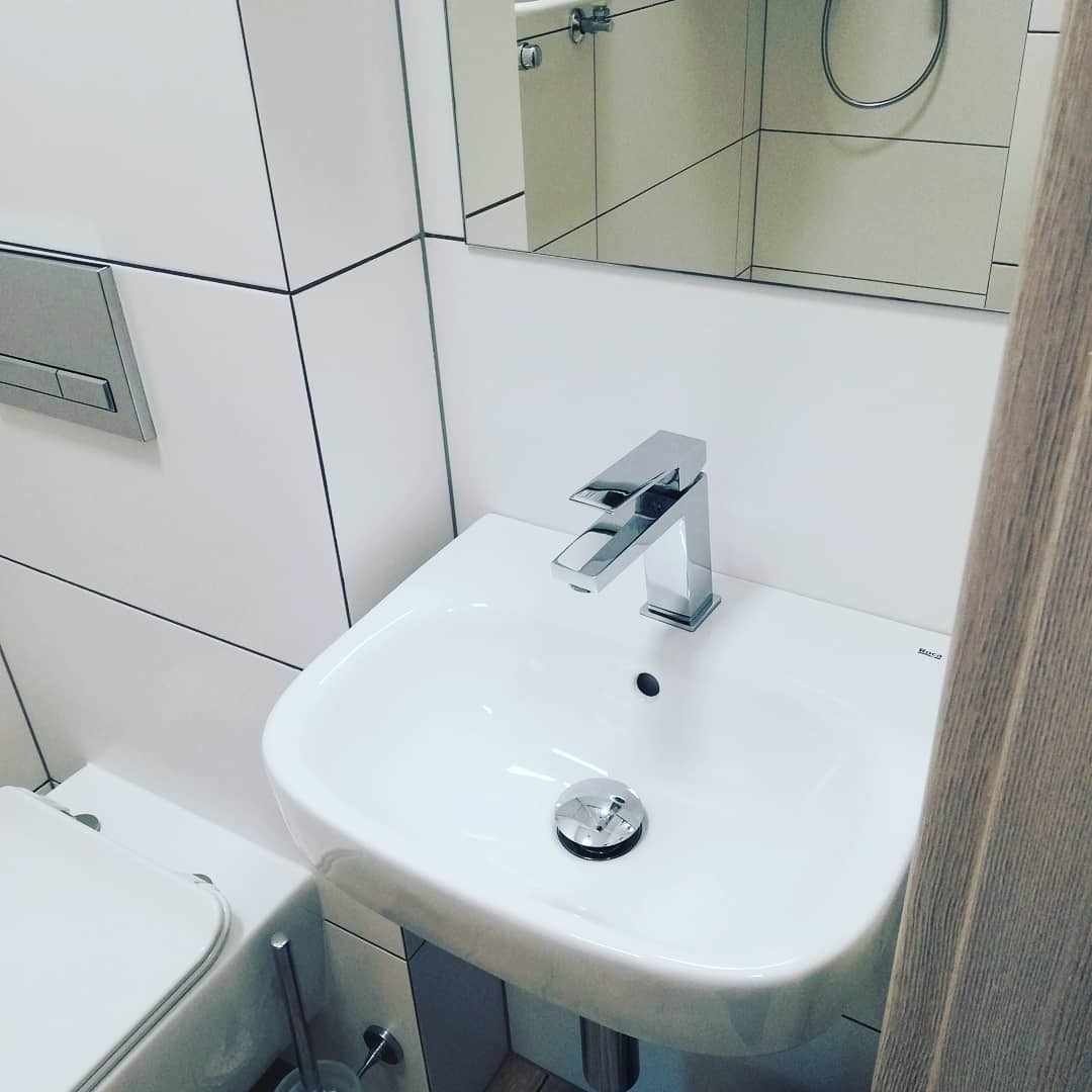 Biała umywalka z chromowaną baterią i odpływem w nowoczesnej łazience z białymi płytkami i lustrem.