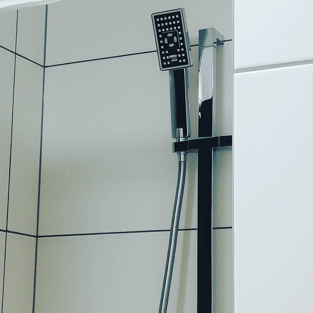 Nowoczesna, kwadratowa słuchawka prysznicowa z chromowanym wykończeniem i czarnym panelem, zamontowana na białej, kafelkowanej ścianie w łazience w Gdyni.