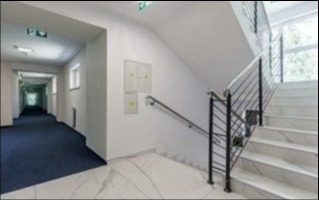 Wnętrze klatki schodowej w budynku biurowym z długim korytarzem w tle. Białe marmurowe schody i podłoga, ciemnoniebieska wykładzina na korytarzu, białe ściany z dwoma skrzynkami elektrycznymi.