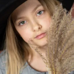 Portret dziewczynki w czarnym kapeluszu, z blond włosami i zielonymi oczami, trzymającej w dłoniach pęk zasuszonej trawy na tle w kolorze pudrowego różu.