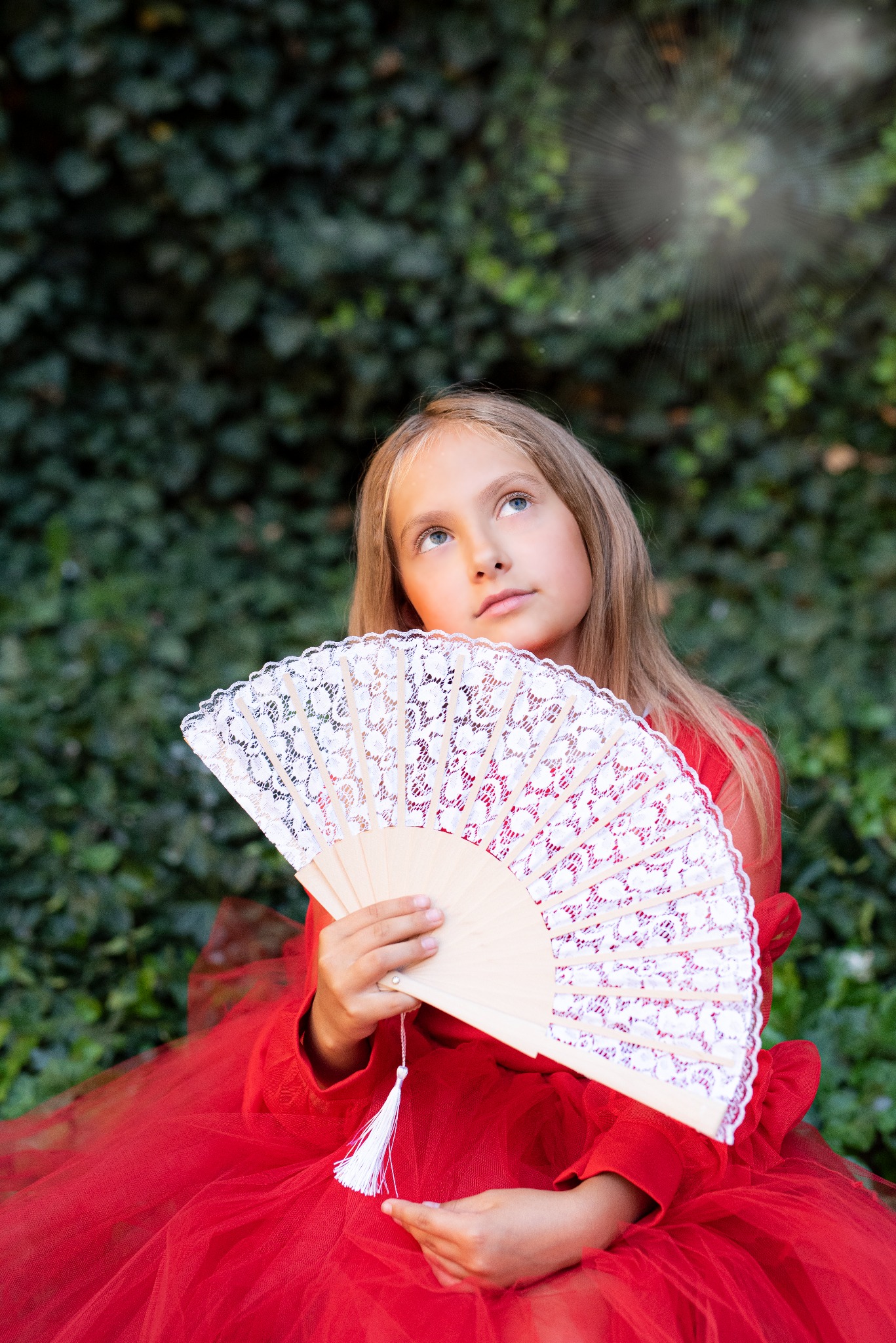 Dziewczynka z blond włosami w czerwonej sukience trzyma koronkowy wachlarz na tle zielonej ściany z bluszczu, spoglądając w górę w kierunku promieni słonecznych.