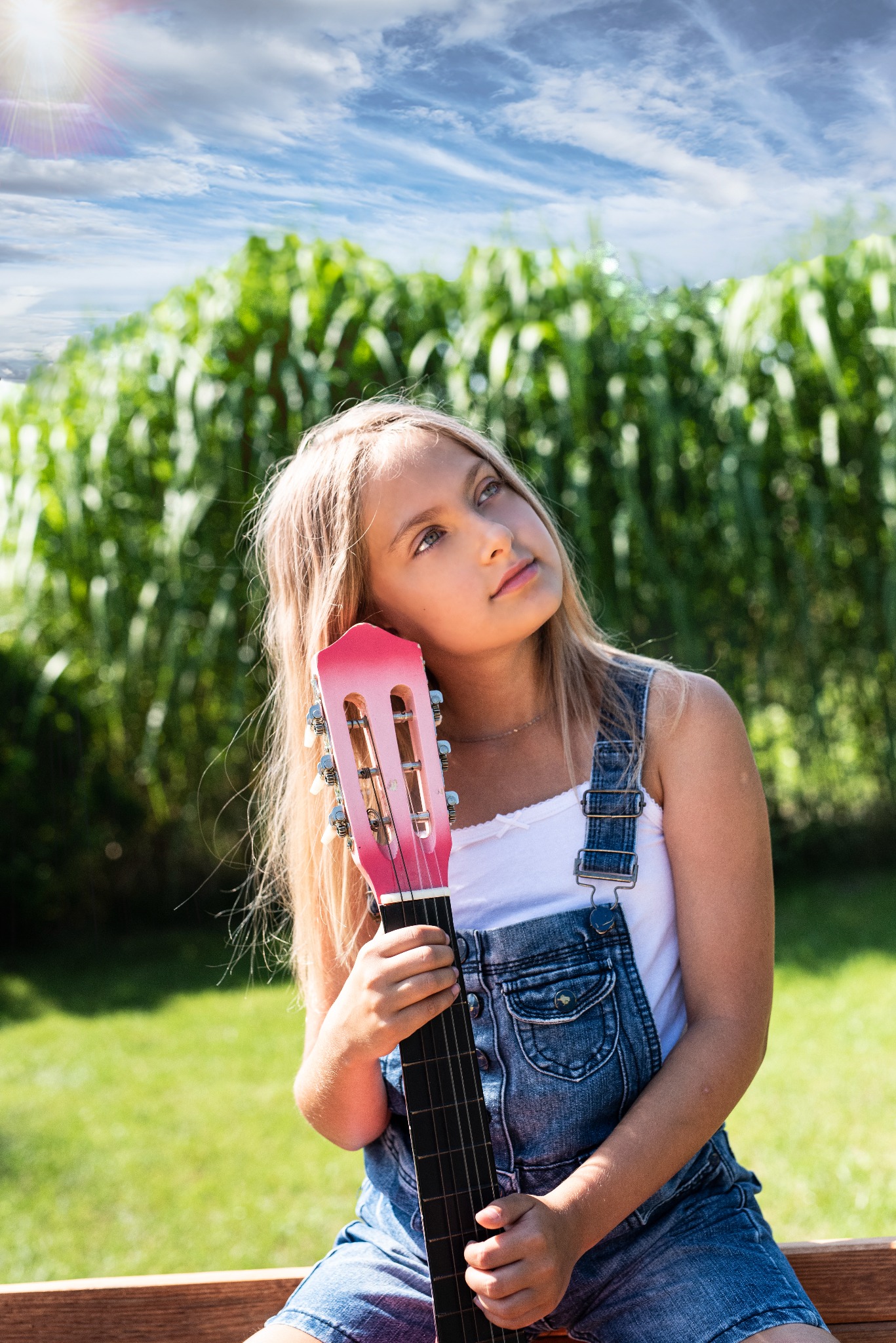 Młoda blondynka w dżinsowych ogrodniczkach opiera głowę o różową gitarę, spoglądając w górę na tle zieleni i błękitnego nieba.
