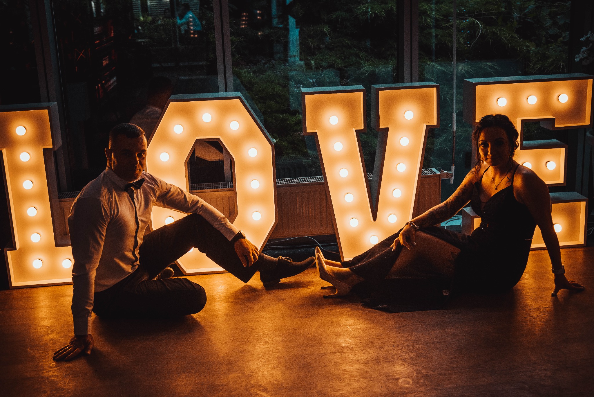 Para siedzi na podłodze obok dużego, podświetlanego napisu LOVE, on w koszuli z muchą, ona w długiej sukni z tatuażem na ramieniu.