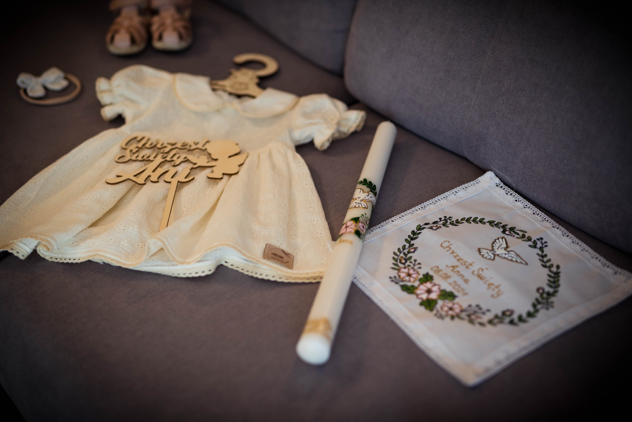 Przygotowania do chrztu: biała sukienka z drewnianym napisem 'Chrzest Święty Ania', świeca z gołębiem i haftowana chusteczka z datą chrztu, leżące na szarej sofie.