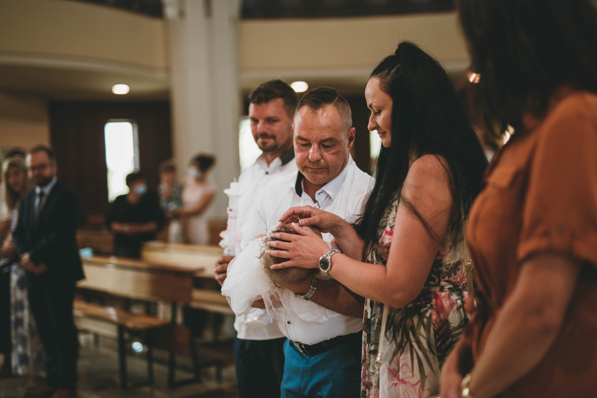 Rodzice i ojciec chrzestny nad dzieckiem podczas ceremonii chrztu w kościele, widoczne ławki i inni goście.