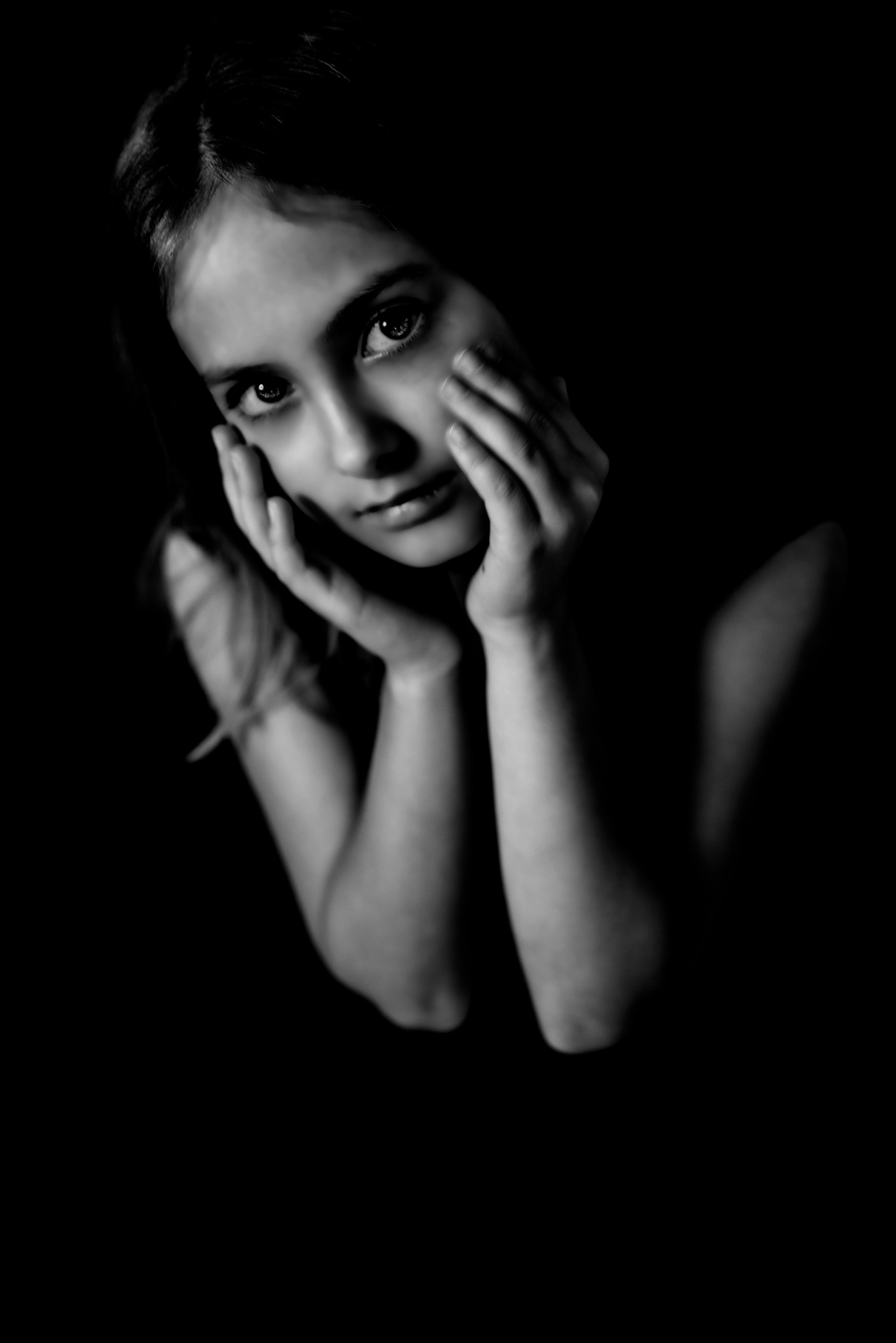 Portret młodej kobiety w czerni i bieli, dłonie podpierają twarz, skupione spojrzenie w obiektyw, silny kontrast światła i cienia.