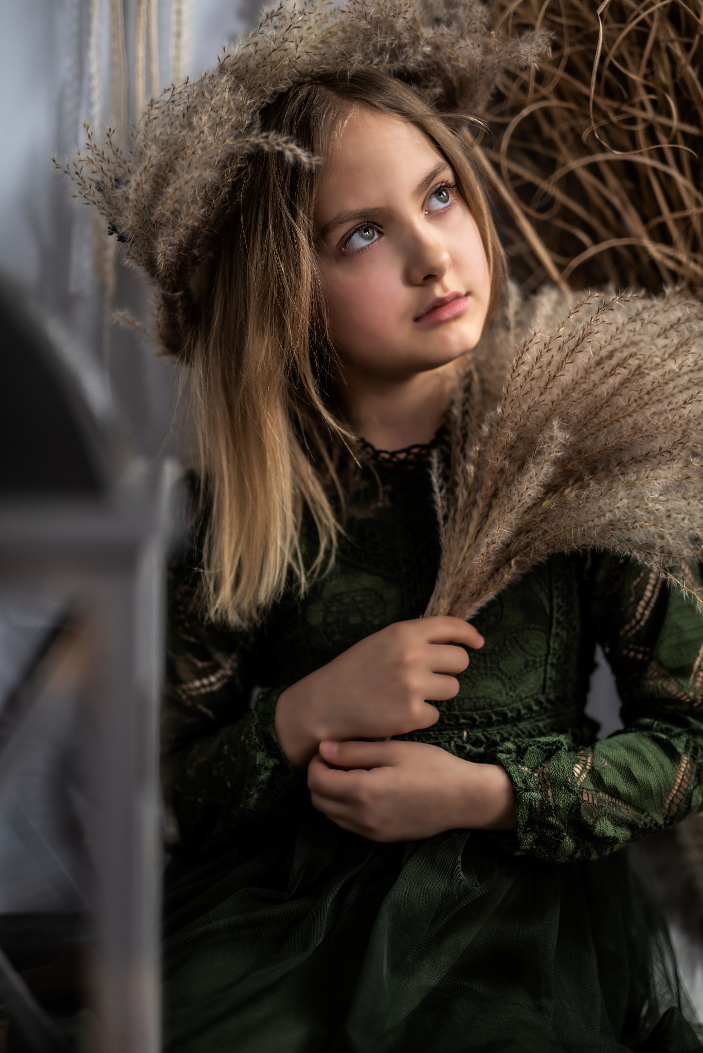 Portret młodej dziewczyny w zielonej sukience, z wiankiem z trawy pampasowej na głowie i bukietem w rękach, spoglądającej w górę w miękkim świetle.