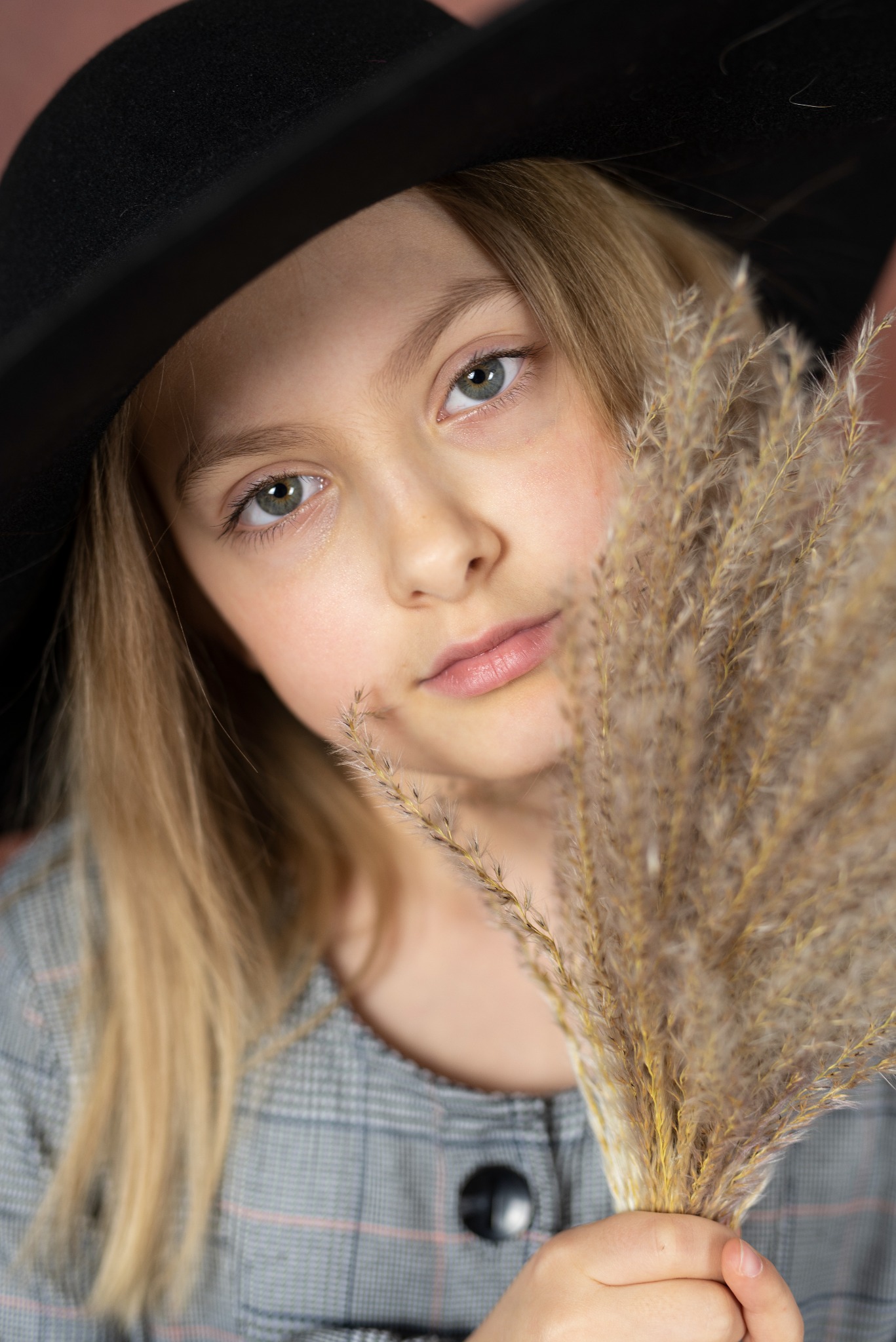Portret dziewczynki w czarnym kapeluszu, z blond włosami i zielonymi oczami, trzymającej w dłoniach pęk zasuszonej trawy na tle w kolorze pudrowego różu.