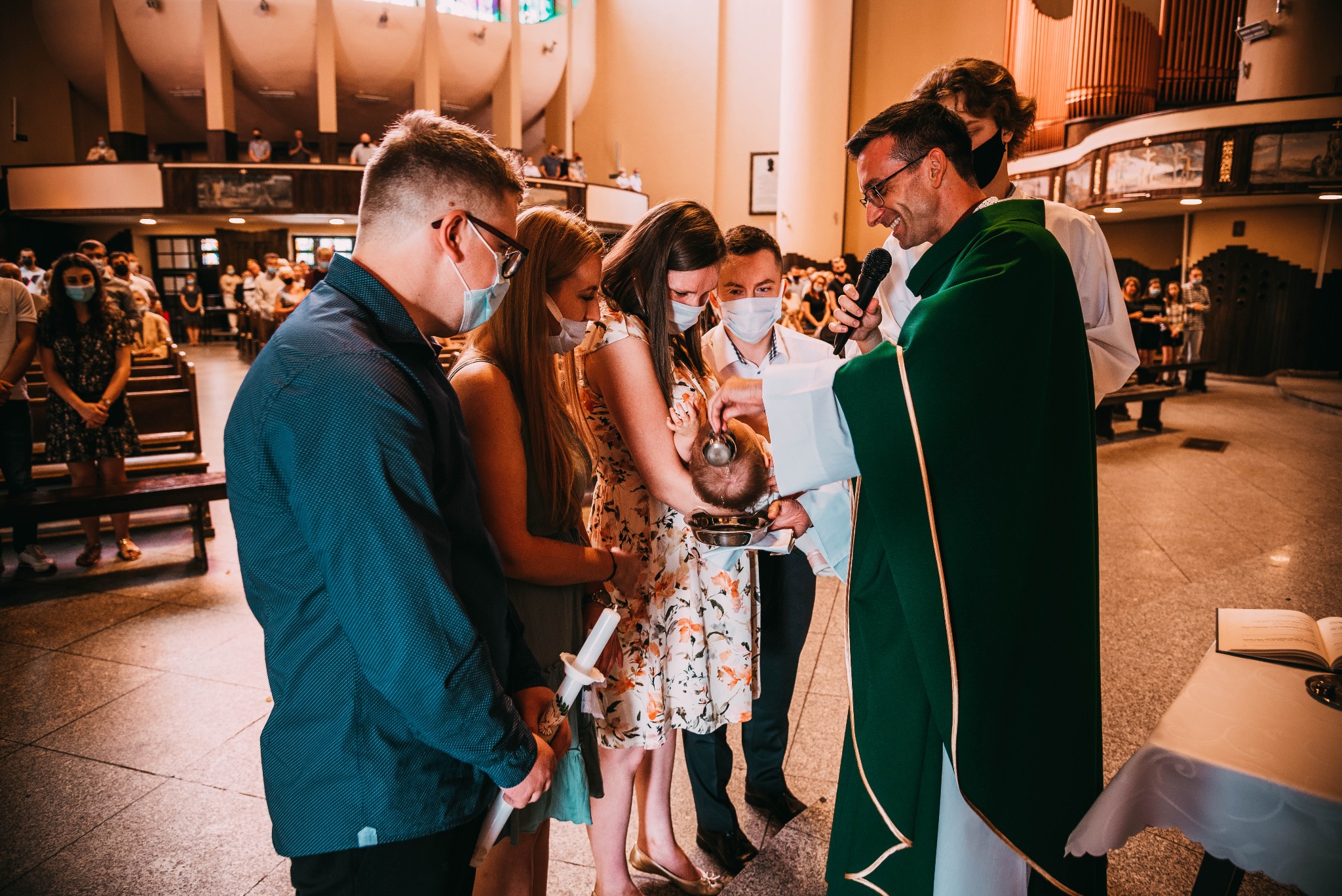 Ksiądz w zielonej szacie udziela chrztu dziecku w kościele, otoczony przez rodzinę w maskach ochronnych, w tle widoczni wierni na ławkach.