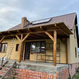 Dom z drewnianą konstrukcją zadaszenia tarasu, drabiny oparta o ścianę, panele słoneczne na brązowym dachu i ceglana podmurówka tarasu.