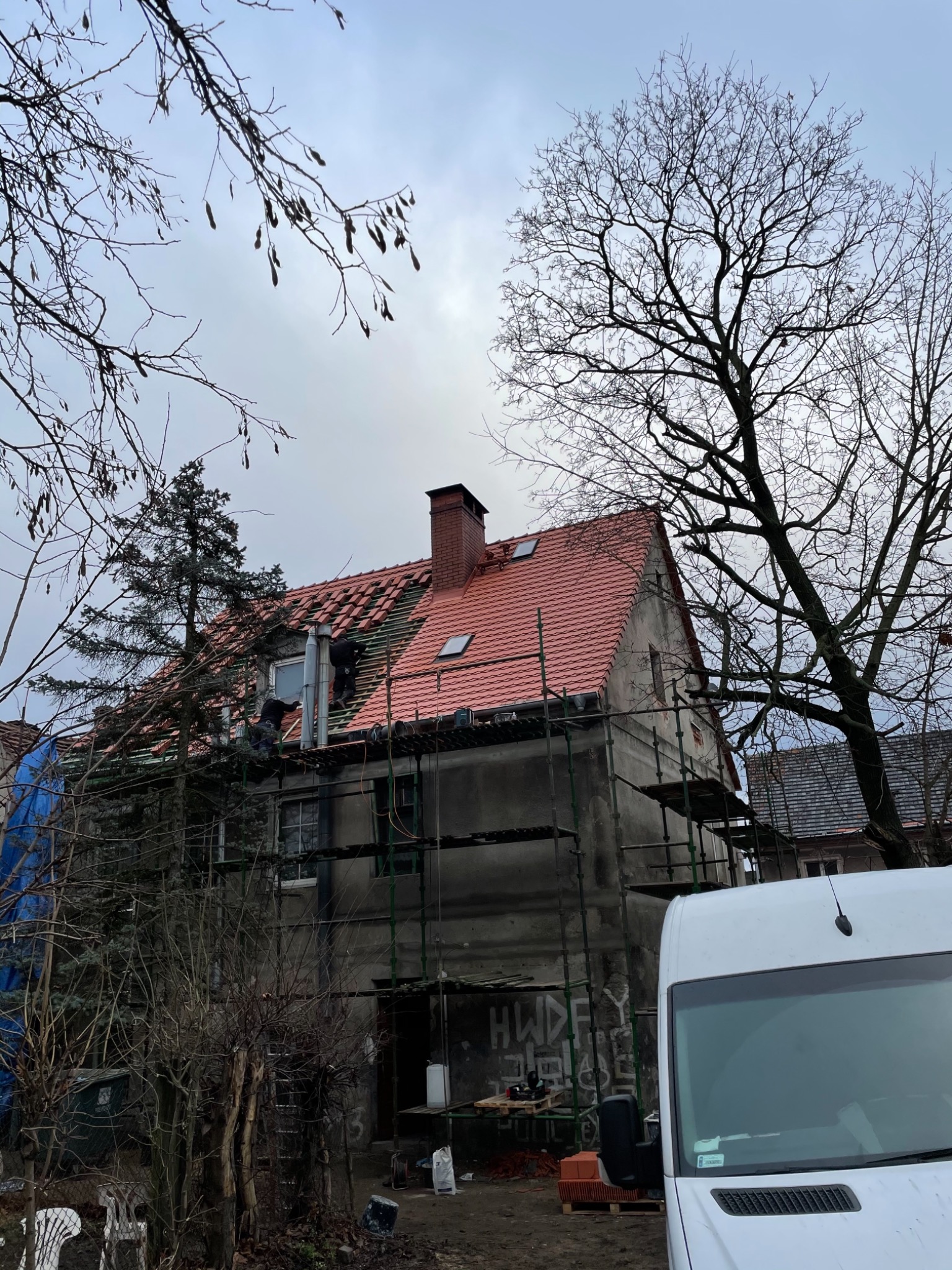 Renowacja dachu z czerwonej dachówki na starym, dwupiętrowym budynku, widoczna z poziomu ulicy, z rusztowaniem i pracownikami na dachu, przysłonięta częściowo drzewami bez liści i zaparkowanym...