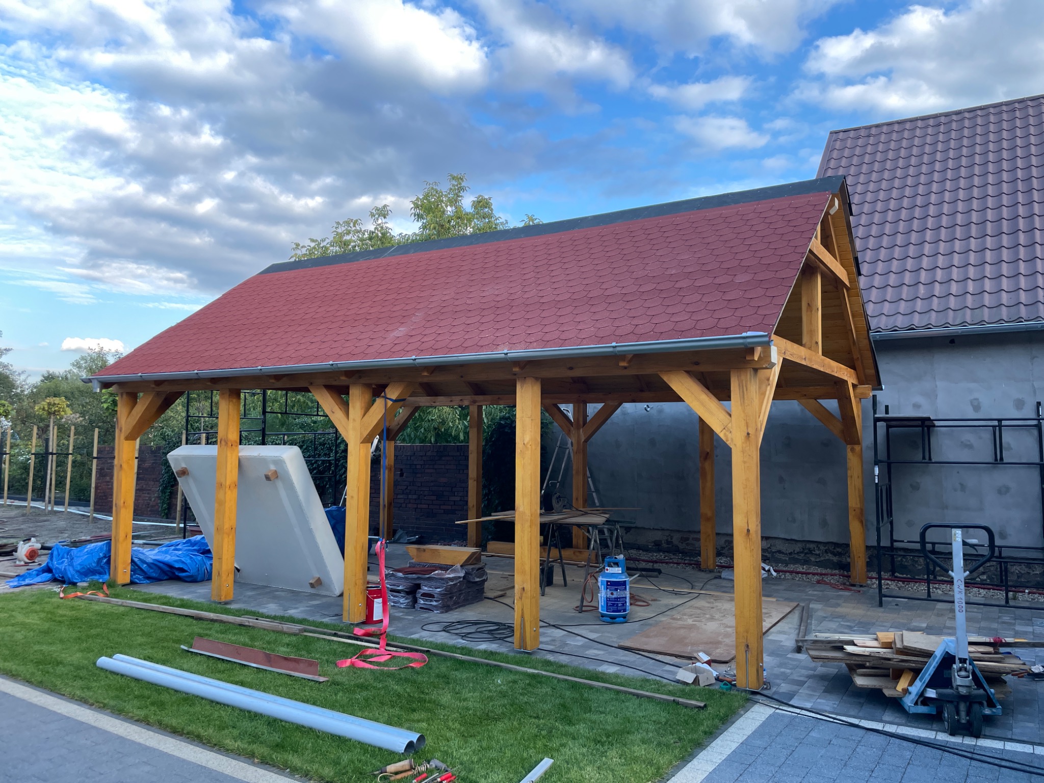Budowa drewnianej altany z czerwoną dachówką, widoczne materiały budowlane, trawa i niebieskie niebo z chmurami.