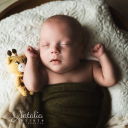 Natalia Bijata Fotografia - Śpiący noworodek owinięty w oliwkowy kocyk, leżący na białej, wzorzystej poduszce w drewnianej misce, obok niego leży szydełkowa żyrafa.