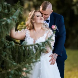 Para młoda w objęciach na tle zieleni. Panna młoda w koronkowej sukni, pan młody w granatowym garniturze. Romantyczna sesja ślubna w plenerze, delikatne światło.