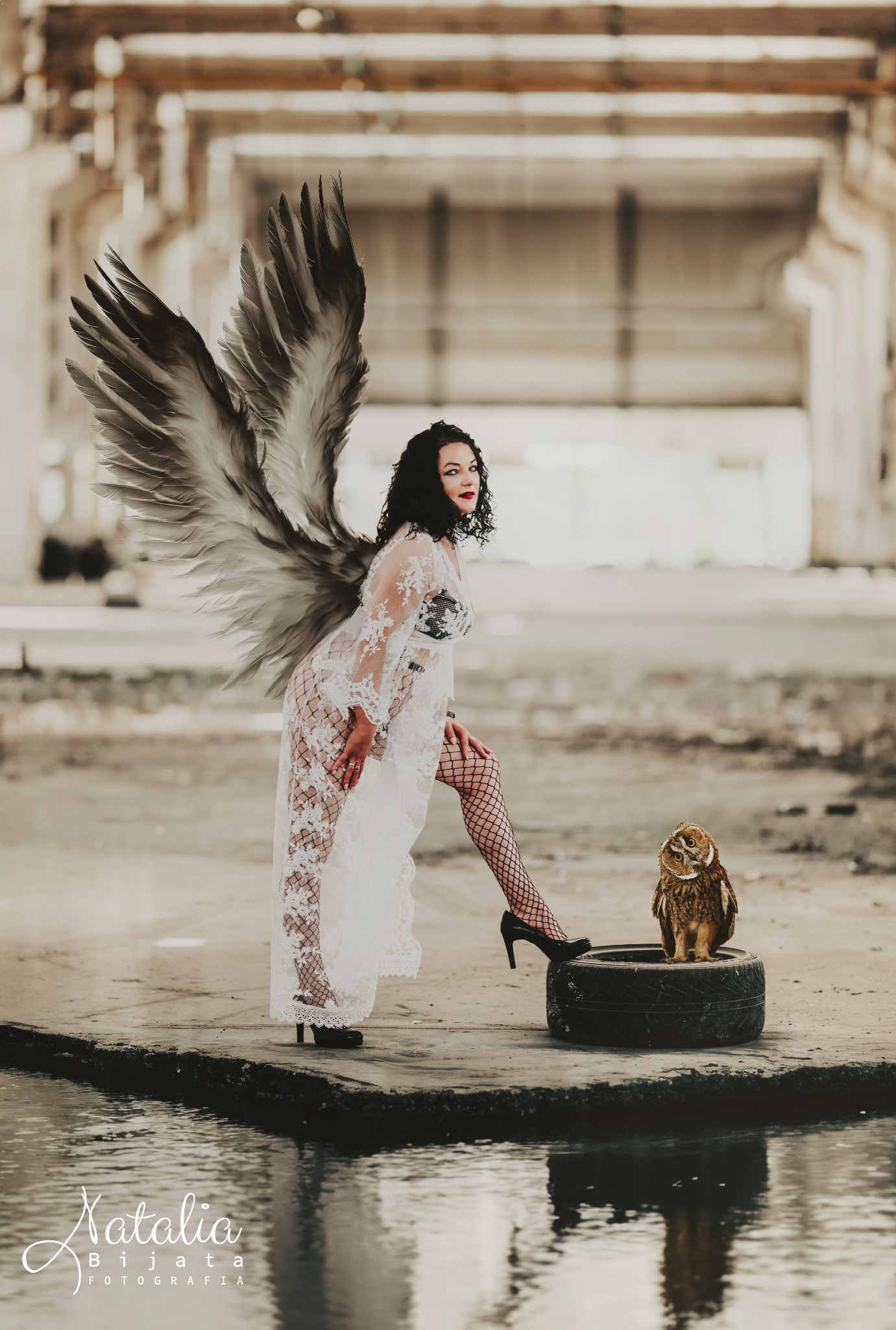 Kobieta w koronkowej sukni i skrzydłach pozuje z sową na oponie w opuszczonym budynku.