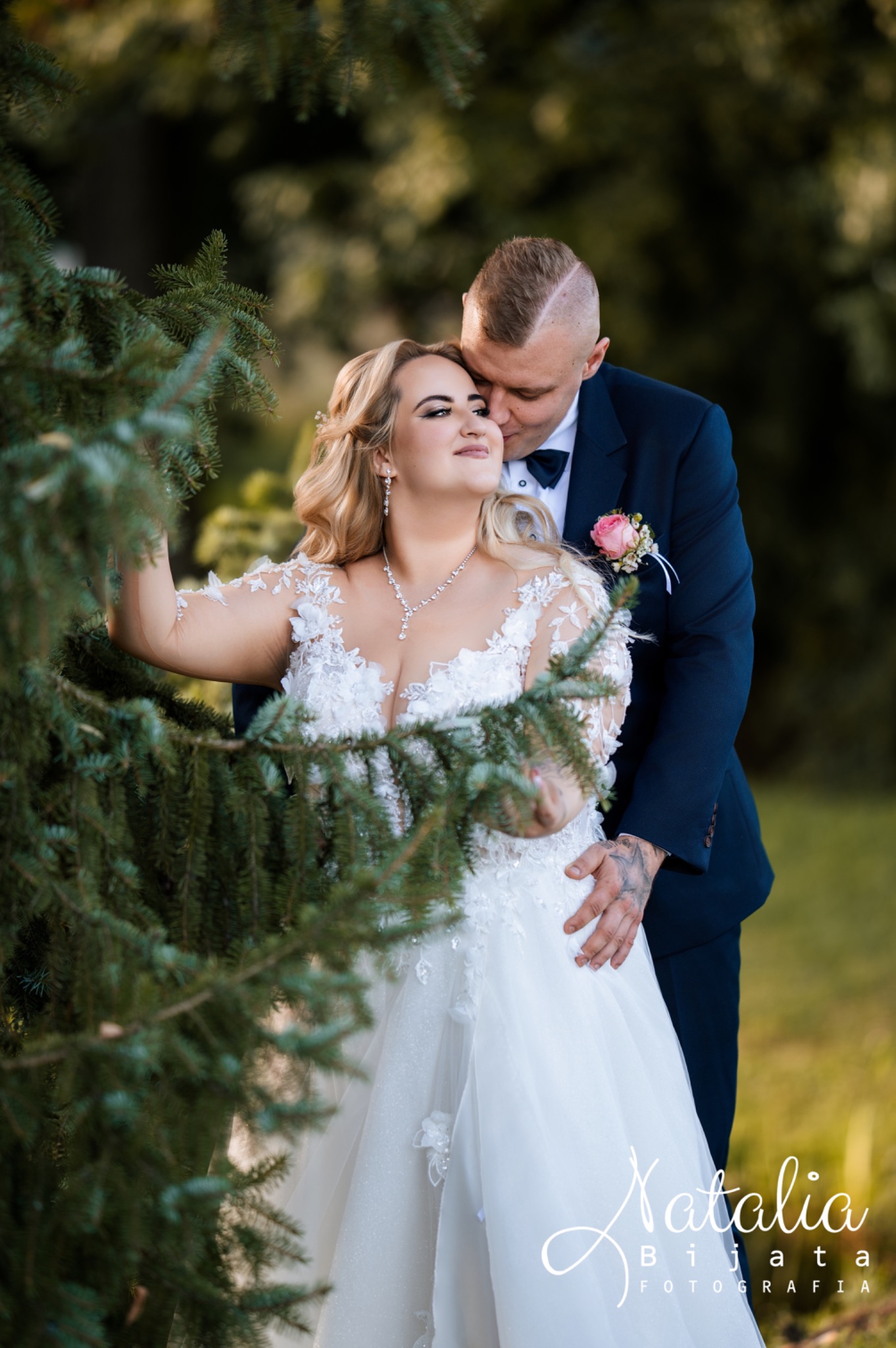 Para młoda w objęciach na tle zieleni. Panna młoda w koronkowej sukni, pan młody w granatowym garniturze. Romantyczna sesja ślubna w plenerze, delikatne światło.