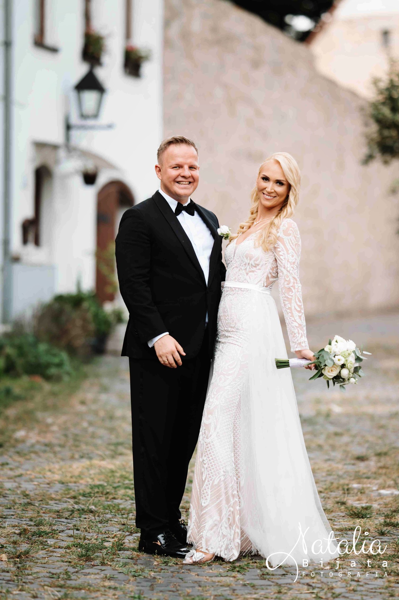 Uśmiechnięta para młoda: pan młody w czarnym smokingu z muchą, panna młoda w koronkowej sukni z bukietem na tle jasnej architektury.