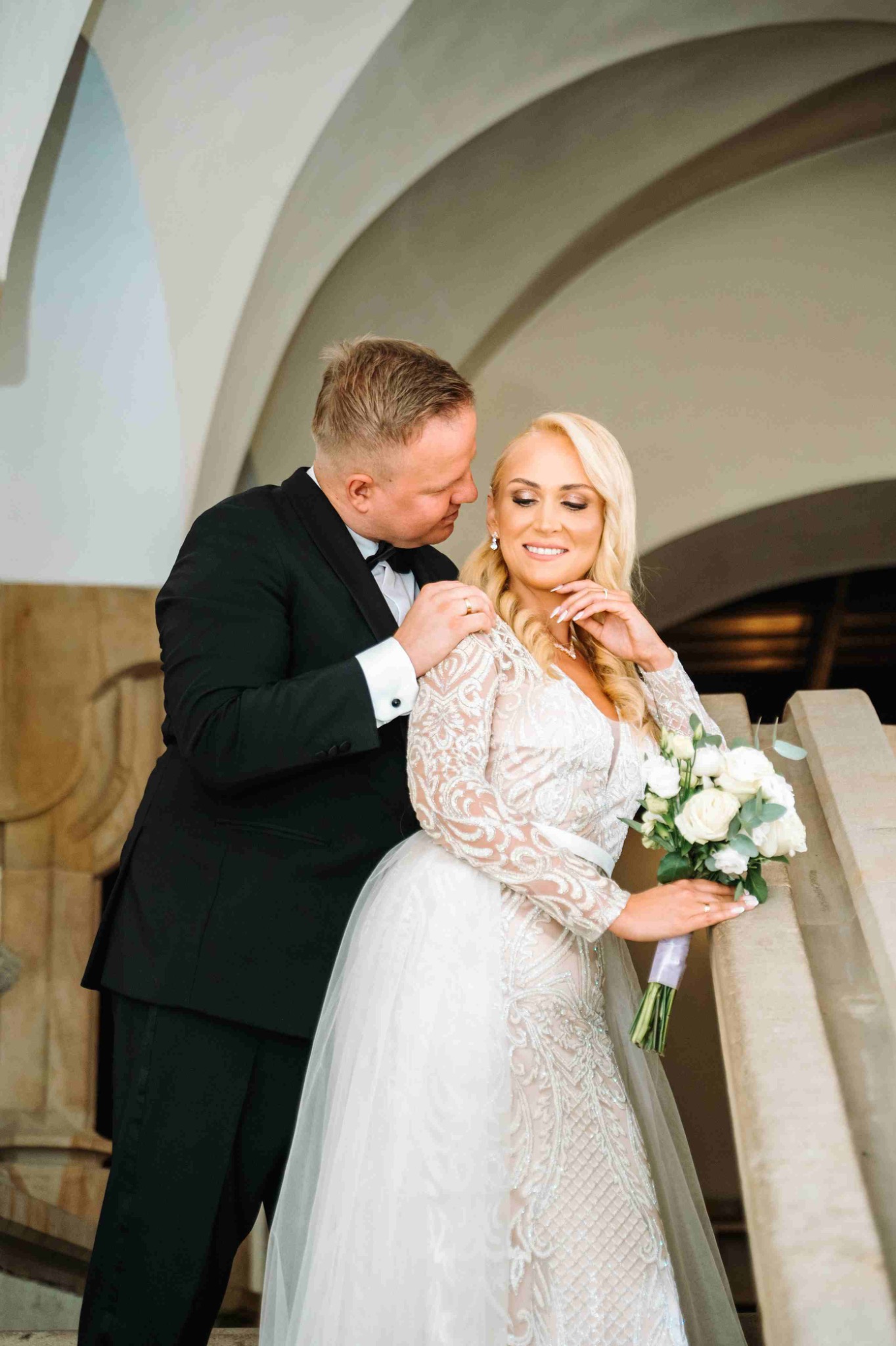 Elegancka para młoda: blond panna młoda w koronkowej sukni z bukietem, pan młody w smokingu, subtelne oświetlenie podkreśla detale stroju i architekturę tła.