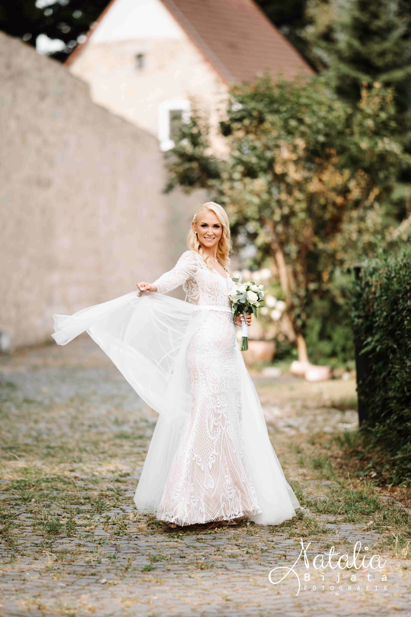 Uśmiechnięta panna młoda w koronkowej sukni z trenem, trzyma bukiet białych róż na tle brukowanej drogi i zieleni. Delikatny welon powiewa na wietrze.