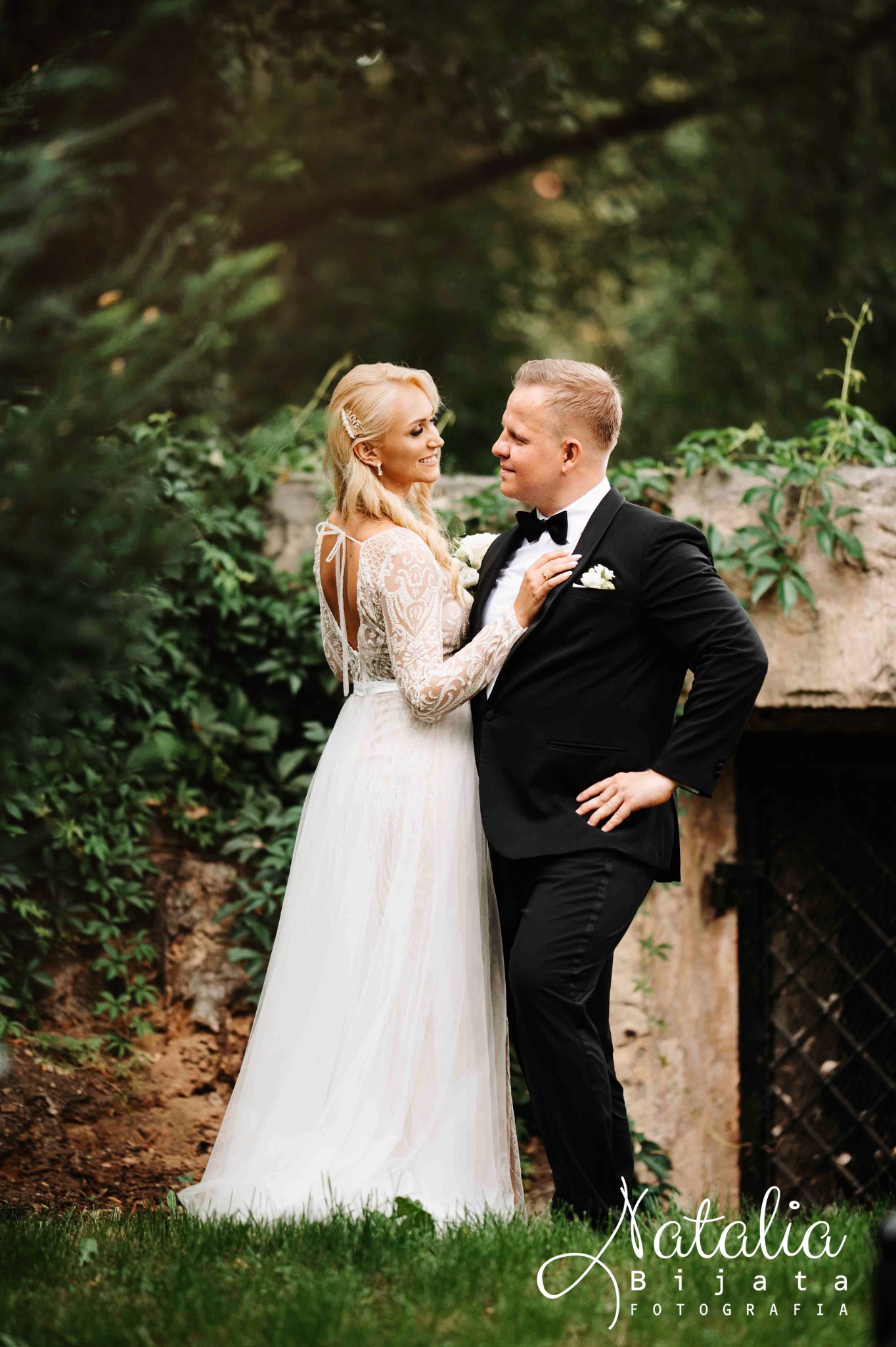 Uśmiechnięta para młoda w plenerze: panna młoda w koronkowej sukni z odkrytymi plecami, pan młody w czarnym smokingu, zielone tło z kamiennym murem.