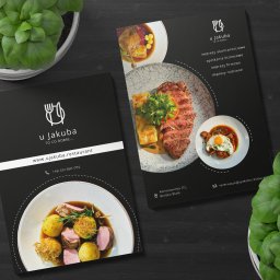 Business Partners Club - Dwa czarne foldery reklamowe restauracji 'u Jakuba' z potrawami, logo i danymi kontaktowymi, leżące na ciemnym drewnianym stole obok doniczek z zielonymi roślinami.