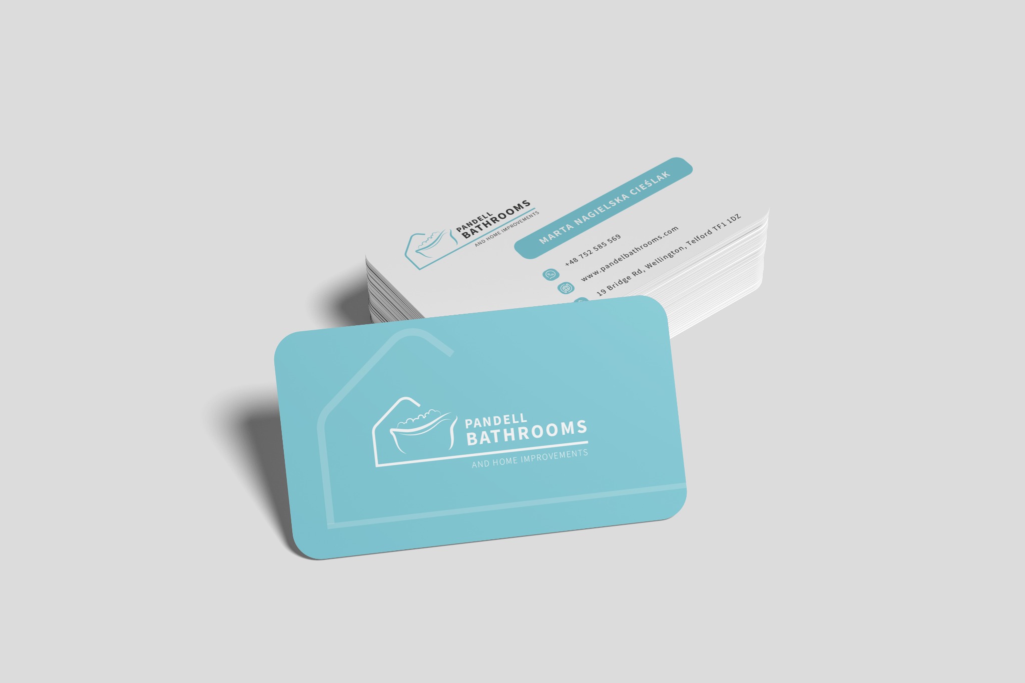 Stos wizytówek firmy remontującej łazienki, z logo przedstawiającym wannę wewnątrz obrysu domu, na jasnoszarym tle.