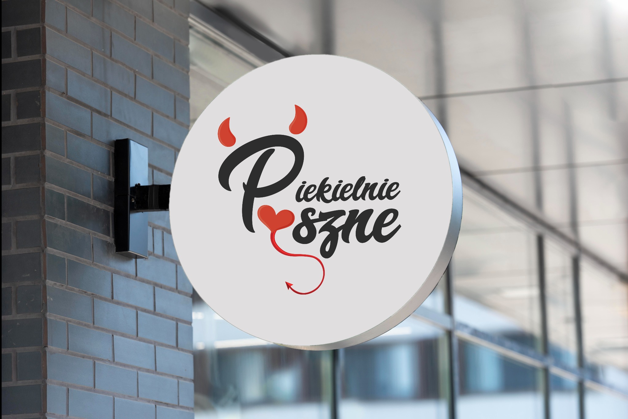 Okrągły szyld reklamowy na ścianie z cegły, z napisem 'Piekielnie Pyszne' w czcionce stylizowanej na pismo odręczne, z elementami graficznymi rogów i ogona diabła w kolorze czerwonym.