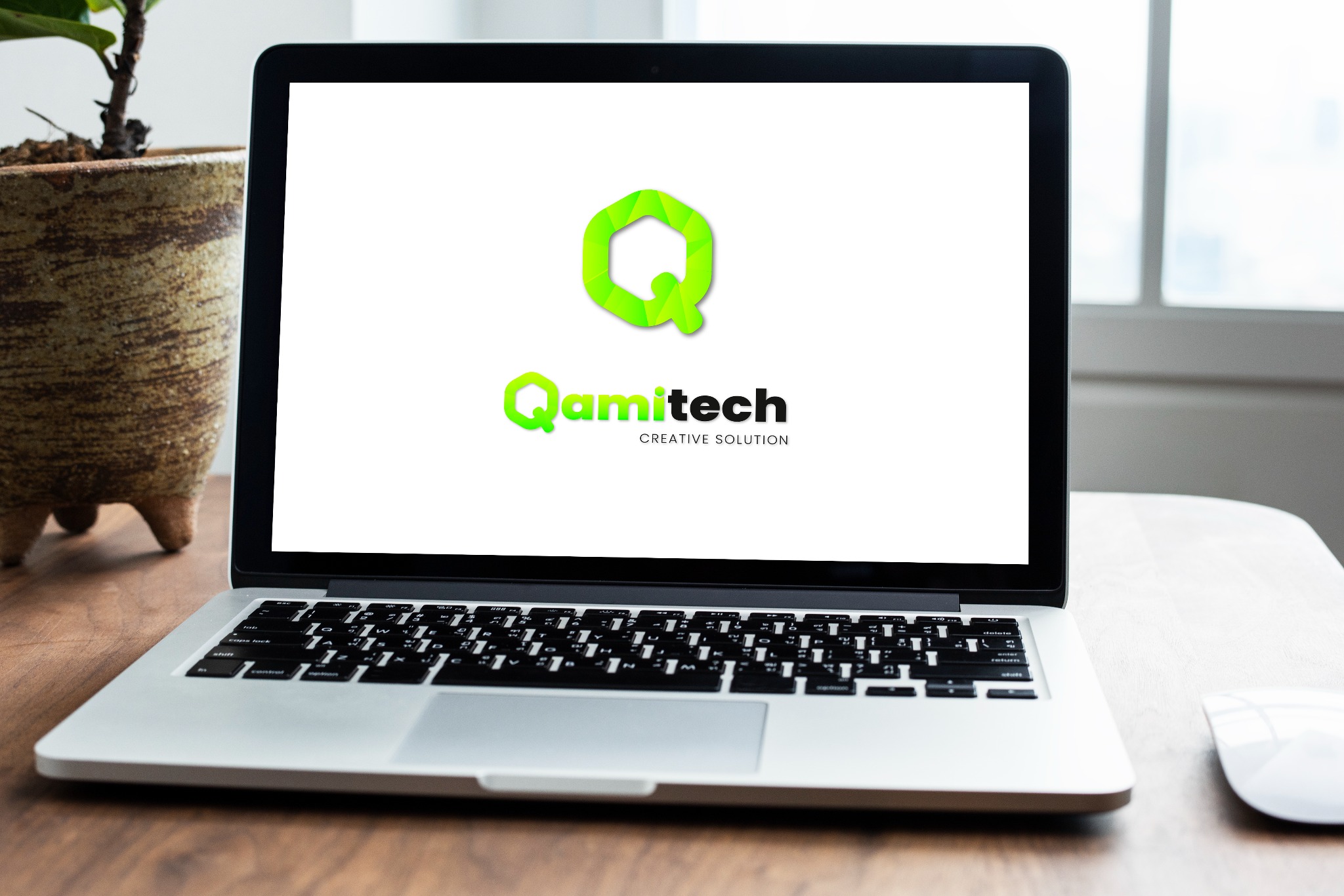 Laptop z wyświetlonym logo firmy Qamitech Creative Solution na ekranie, obok doniczka z rośliną i biała myszka na drewnianym biurku.