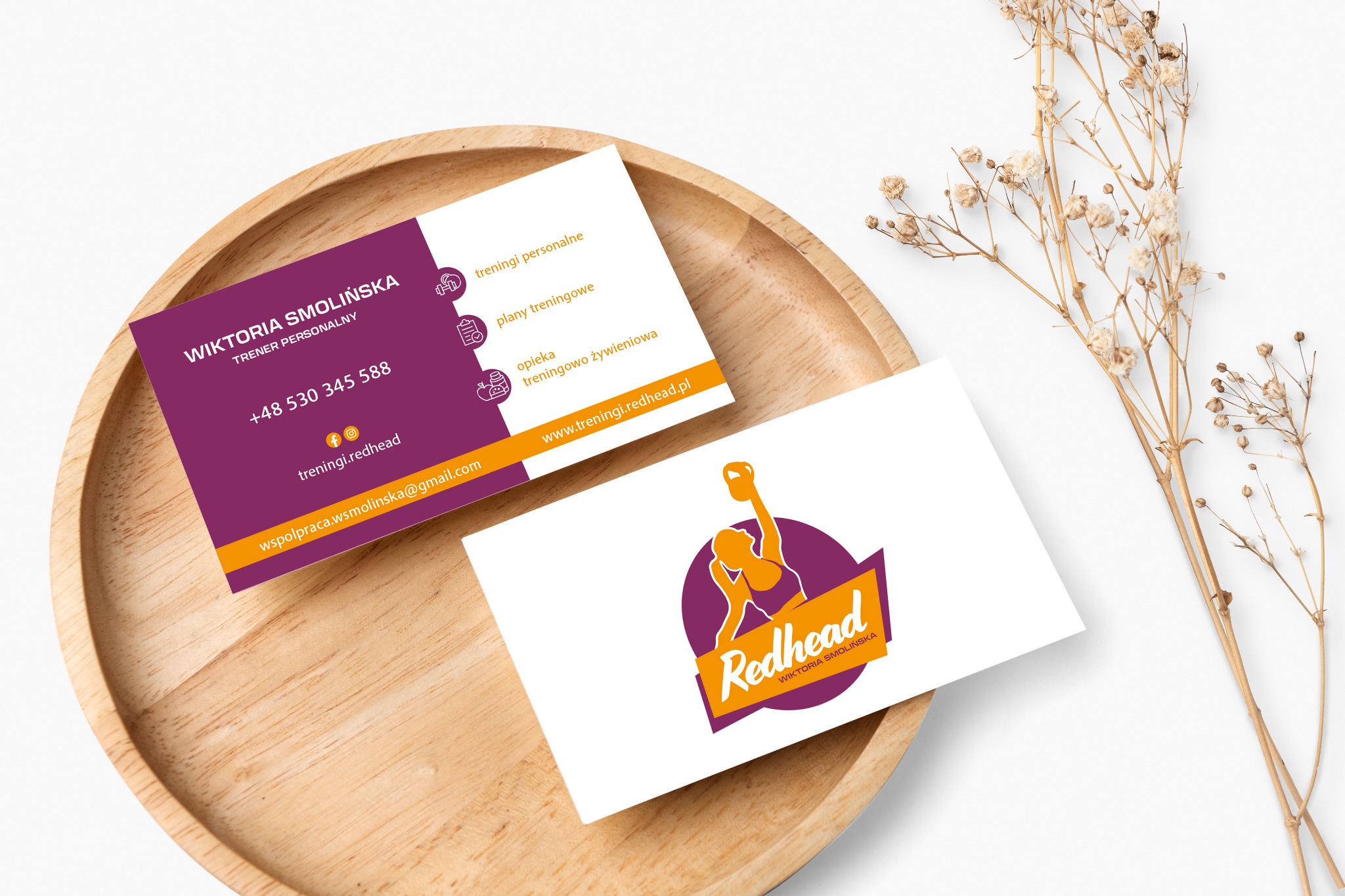 Dwie wizytówki na drewnianej tacy z suszonymi kwiatami. Jedna wizytówka w kolorze fioletowym i złotym zawiera dane kontaktowe trenera personalnego, druga wizytówka z logo firmy treningowej...