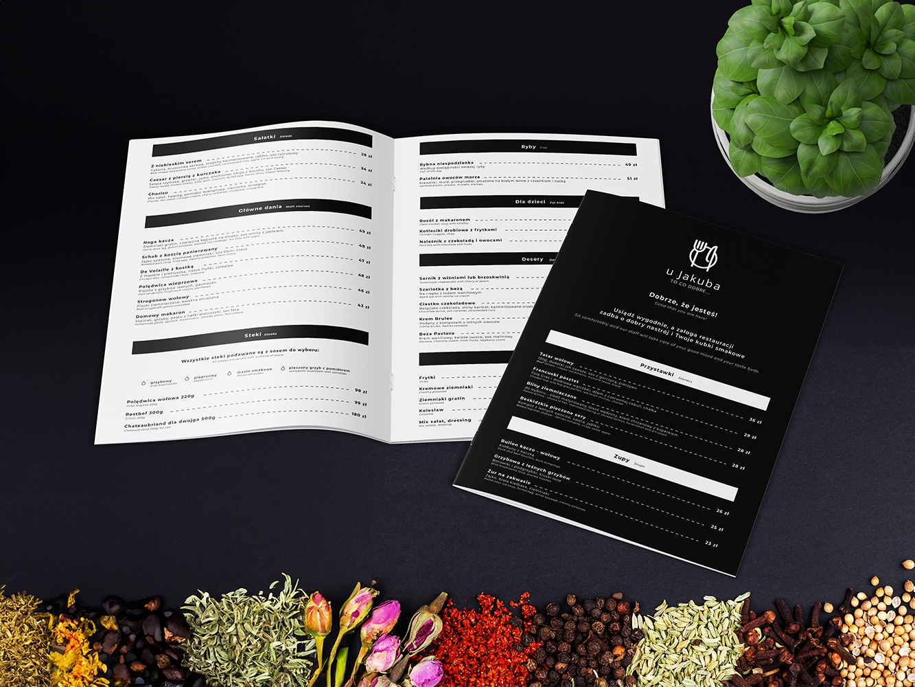 Dwa rozłożone menu restauracji U Jakuba na czarnym tle, otwarte na stronach z sałatkami, daniami głównymi, stekami, rybami, deserami i przystawkami, otoczone przyprawami i ziołami, z doniczką...