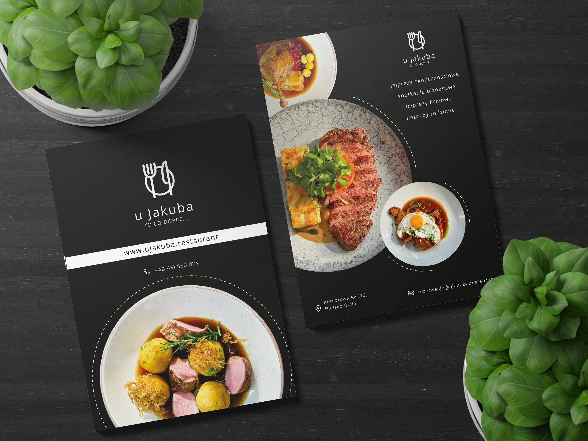 Dwa czarne foldery reklamowe restauracji 'u Jakuba' z potrawami, logo i danymi kontaktowymi, leżące na ciemnym drewnianym stole obok doniczek z zielonymi roślinami.