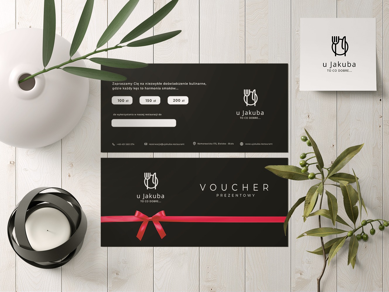 Dwa czarne vouchery prezentowe restauracji 'U Jakuba' z czerwonymi wstążkami, biała wizytówka z logo, ceramiczny wazon i świecznik na tle drewnianych desek.