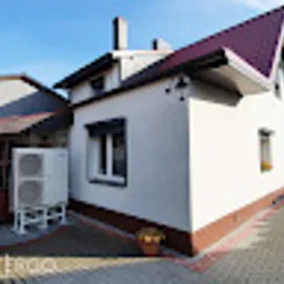 Biały, dwukondygnacyjny dom z brązowym dachem i jasną elewacją, na zewnątrz zainstalowana dwuczęściowa pompa ciepła powietrze-woda, widoczna z boku, na tle kostki brukowej.