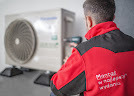 Mężczyzna w czerwonej kurtce z logo firmy montuje klimatyzator na ścianie, używając wkrętarki akumulatorowej. Widoczna jednostka zewnętrzna klimatyzacji, częściowo zasłonięta przez sylwetkę montera.