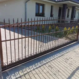 Przesuwna brama metalowa w kolorze brązowym z ozdobnymi elementami, zamontowana na posesji z kostki brukowej, rzucająca charakterystyczny cień na podjazd.