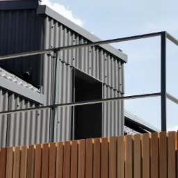 Nowoczesna balustrada ze stali malowanej proszkowo na tle falistej blachy elewacyjnej i drewnianego ogrodzenia z pionowych desek.