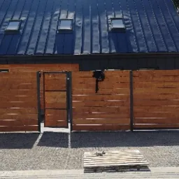 Nowoczesne ogrodzenie z drewnianych paneli i metalowych słupków przed domem z ciemnym dachem i trzema oknami dachowymi; dwóch mężczyzn stoi obok ogrodzenia, a na ziemi leżą drewniane palety.